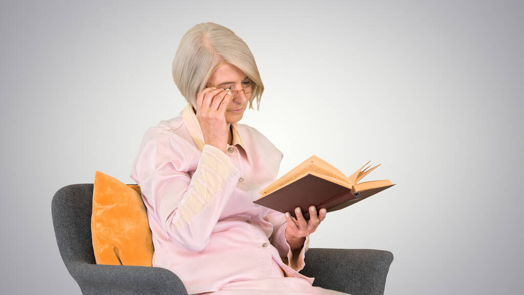 Συνταξιούχος γυναίκα διαβάζει ένα βιβλίο κάθεται σε μια καρέκλα στην κλίση προς τα πίσω - Φωτογραφία, εικόνα