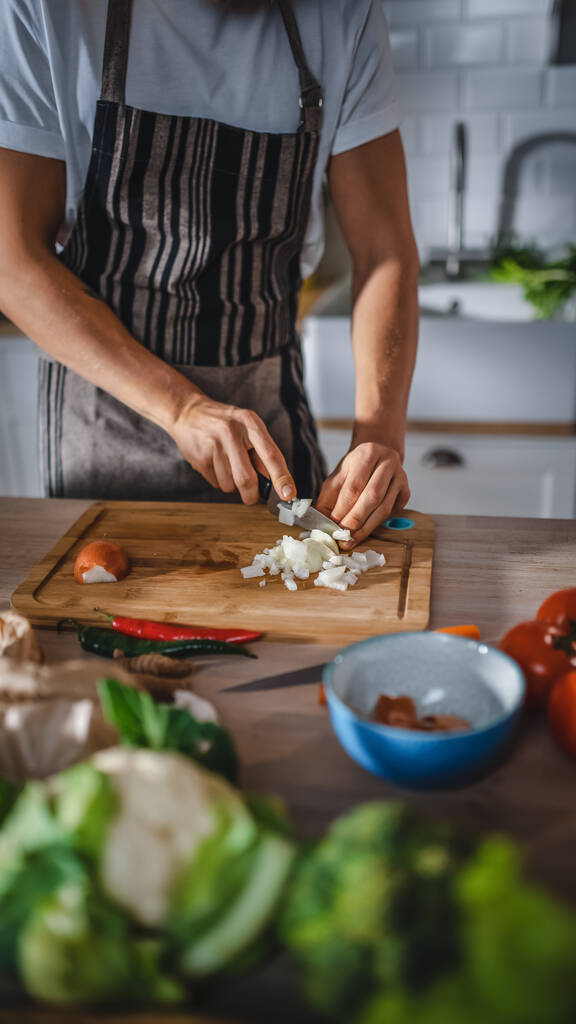 Pohledný muž krájí zelenou jarní cibuli ostrým kuchyňským nožem. Muž v bílé košili a zástěře dělá bio salát jídlo v moderní slunné kuchyni. Přírodní čisté stravy a zdravý životní styl. - Fotografie, Obrázek