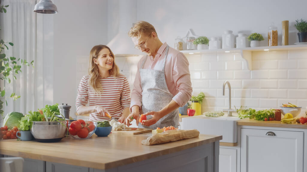Schöner junger Mann in Brille, Schürze und hübsches Mädchen bereiten in der Küche einen Salat zu. Glückliche Paare unterhalten sich beiläufig. Natürliche saubere Ernährung und gesunder Lebensstil. - Foto, Bild