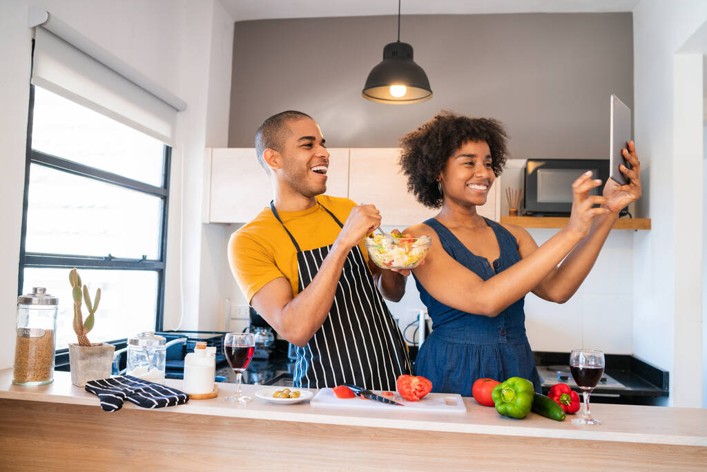 Retrato de pareja latina joven cocinando juntos y tomando una selfie con tableta digital en la cocina en casa. Concepto de relación, cocinero y estilo de vida. - Foto, Imagen
