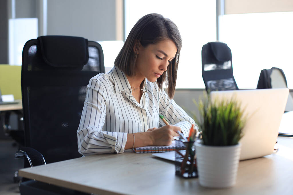 美しいビジネス女性がオフィスの机に座っている間に何かを書いている. - 写真・画像
