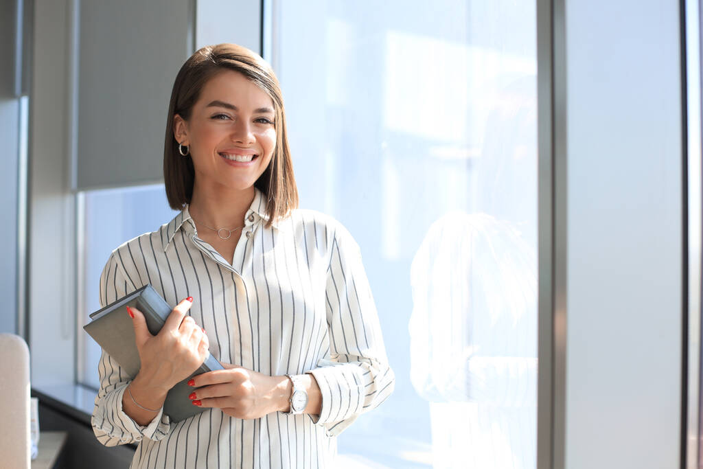 Atrakcyjna kobieta biznesu patrzy w obiektyw i uśmiecha się stojąc w biurze - Zdjęcie, obraz