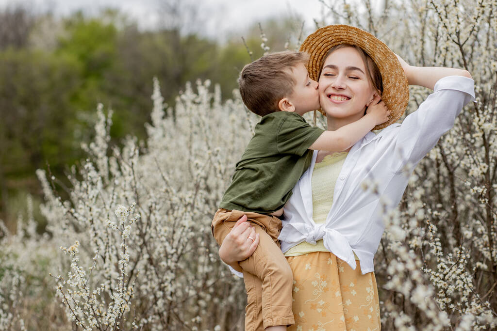 Onnellinen äiti ja poika pitää hauskaa yhdessä. Äiti halaa poikaansa hellästi. Taustalla valkoiset kukat kukkivat. Äitienpäivä. - Valokuva, kuva
