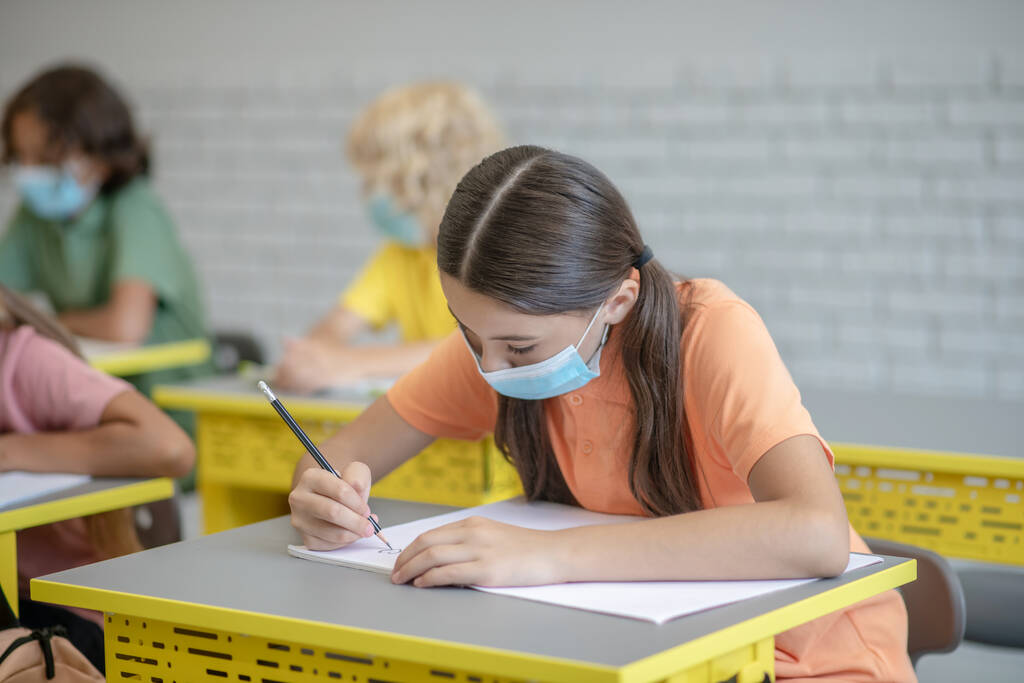 Chica con máscara escribiendo en un cuaderno y buscando involucrado - Foto, imagen