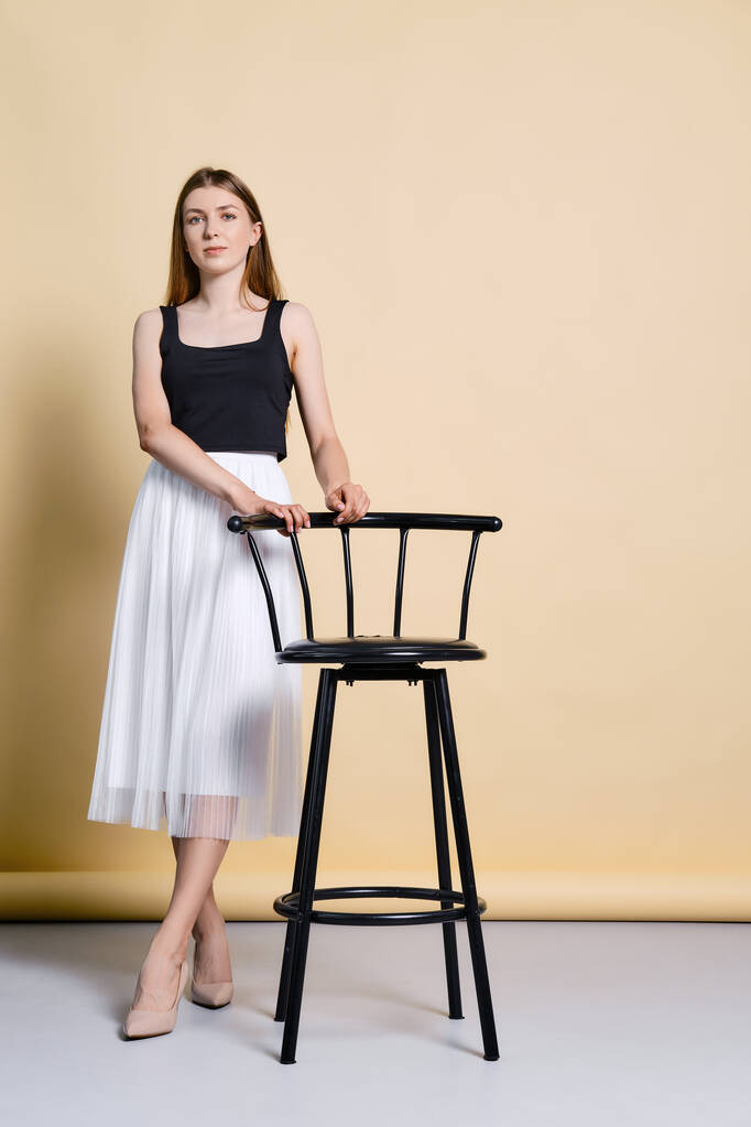 Csinos fiatal nő fekete tartály felső és fehér szoknya áll közel magas szék felett halványsárga háttér - Fotó, kép