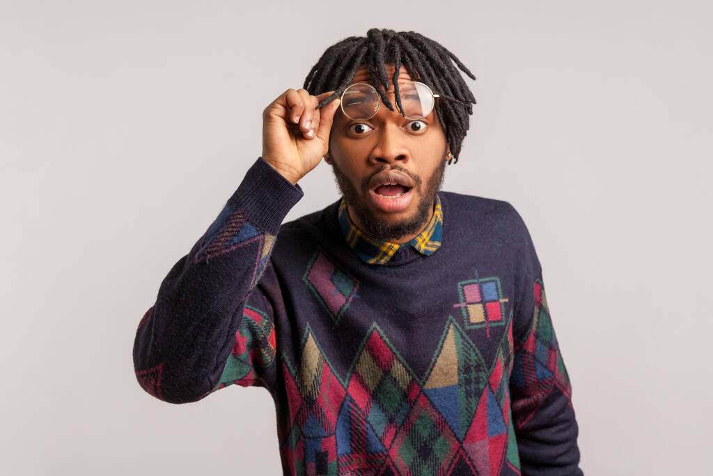 Extremely shocked wondered african man with dreadlocks looking at camera with widely open mouth and big eyes, putting off glasses, surprised. Indoor studio shot isolated on gray background - Photo, Image