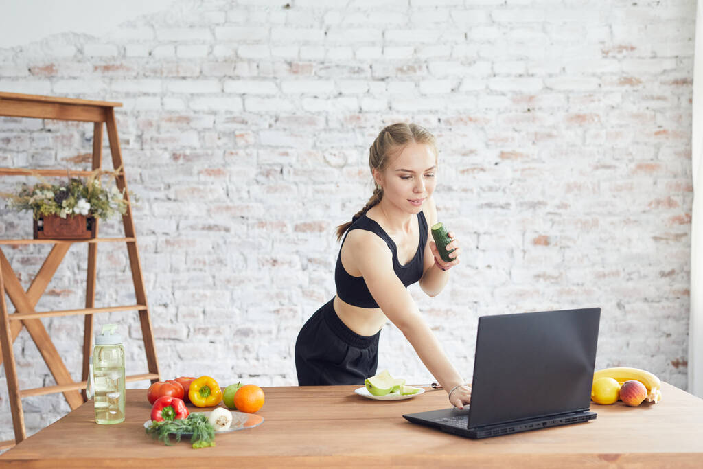 Mooie jonge vrouw draagt sportieve kleding met behulp van laptop, op zoek naar eenvoudige recepten thuis. - Foto, afbeelding