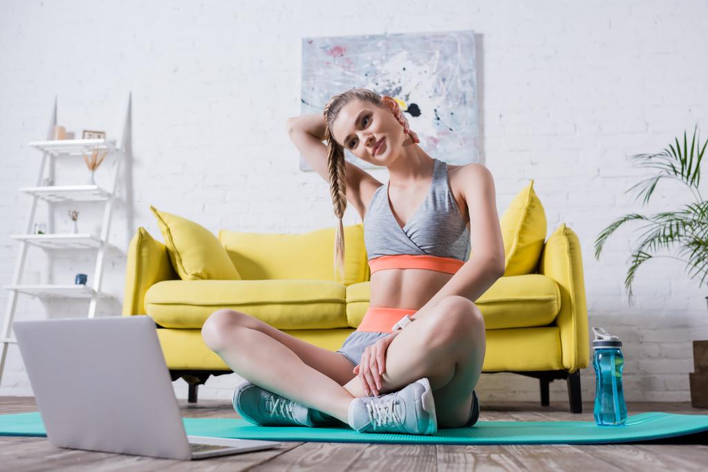 Sportlerin mit Hand hinter dem Kopf streckt sich neben Laptop auf Fitnessmatte - Foto, Bild
