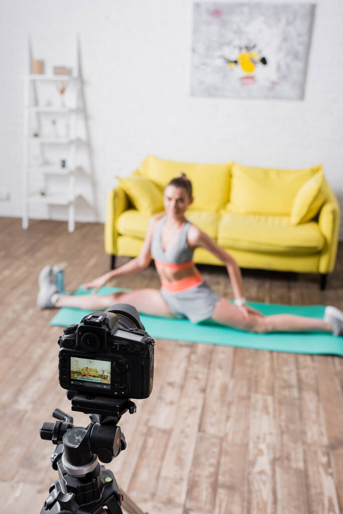 Foco seletivo da jovem esportista fazendo split no tapete de fitness perto da câmera digital em casa  - Foto, Imagem
