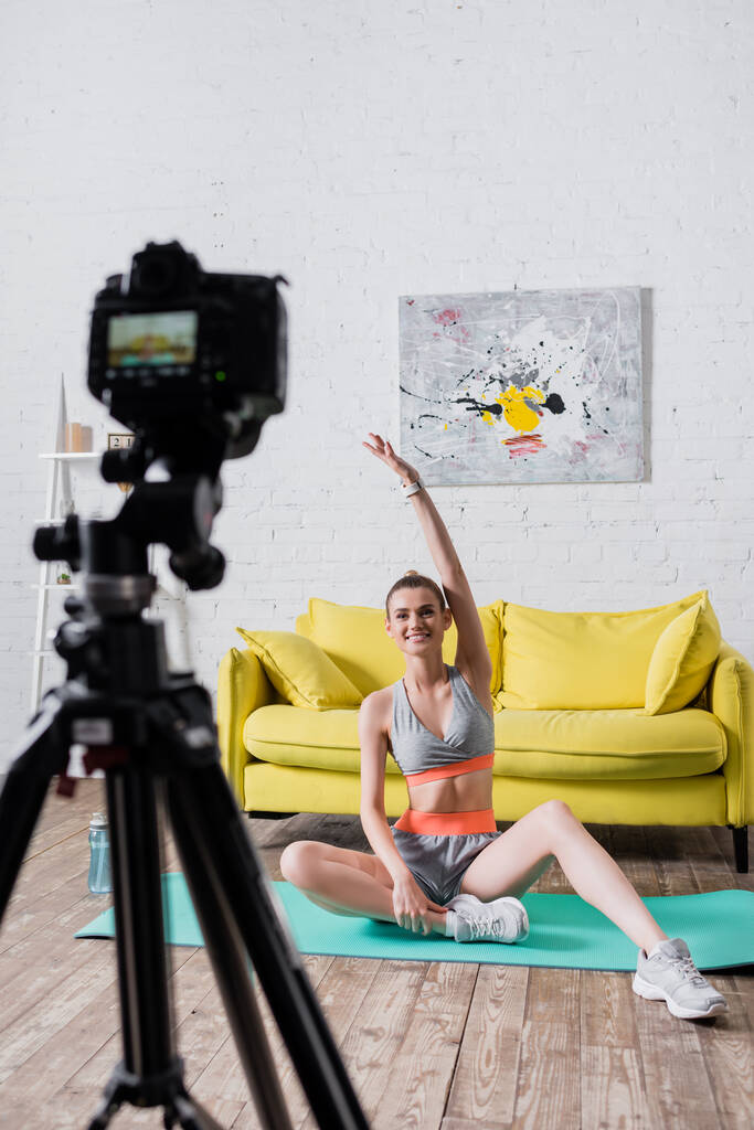 Selectieve focus van lachende sportvrouw die traint op een fitnessmat in de buurt van een digitale camera  - Foto, afbeelding