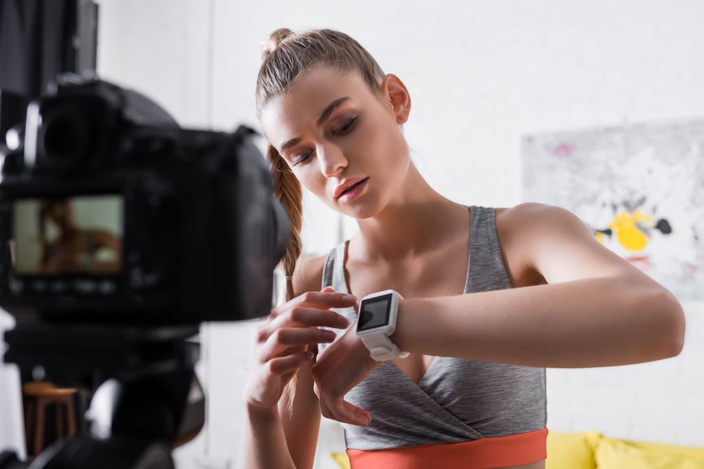 Foco seletivo da mulher em roupas esportivas apontando para o rastreador de fitness perto da câmera digital em casa  - Foto, Imagem