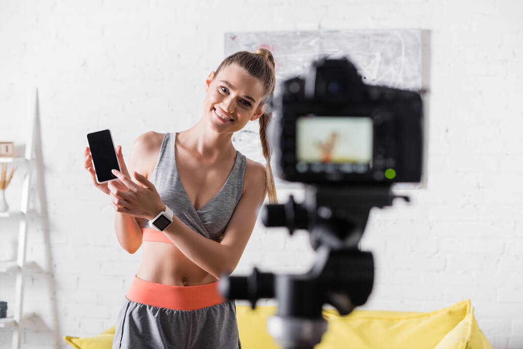 Επιλεκτική εστίαση θετικής αθλήτριας που δείχνει το smartphone κοντά στην ψηφιακή κάμερα στο σπίτι  - Φωτογραφία, εικόνα