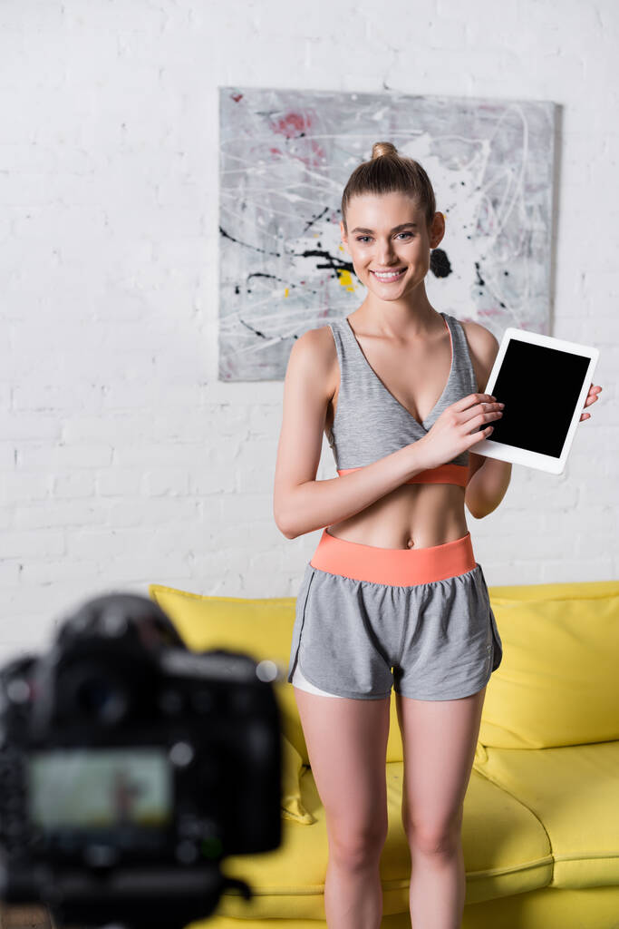 Selectieve focus van lachende sportvrouw met digitale tablet met leeg scherm in de buurt van digitale camera  - Foto, afbeelding