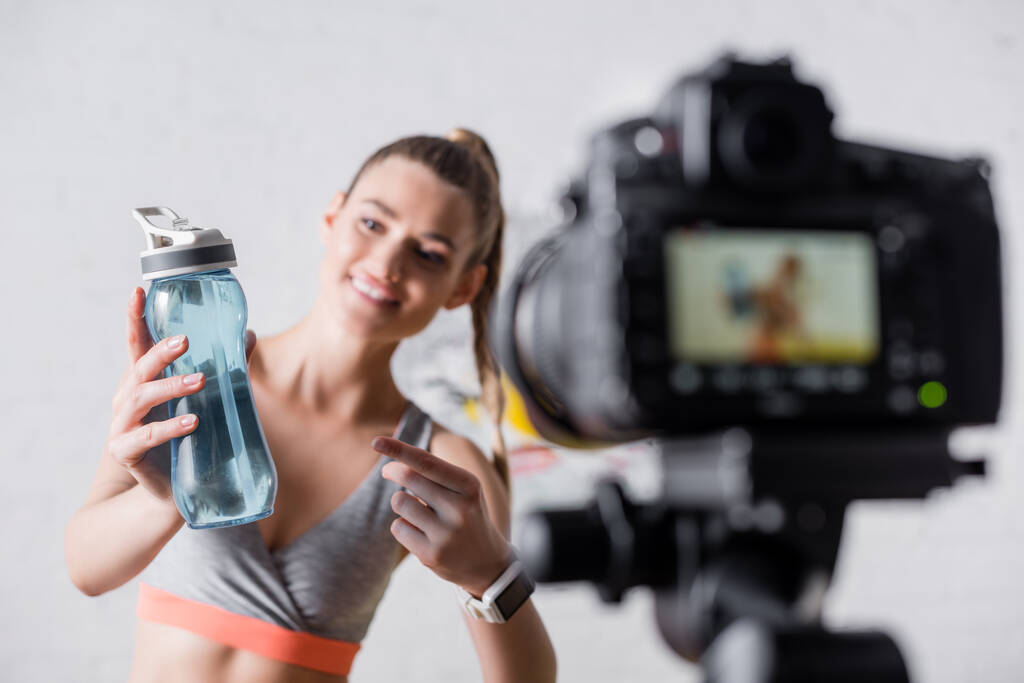 Selectieve focus van vrolijke sportvrouw gericht op sportfles in de buurt van digitale camera thuis  - Foto, afbeelding
