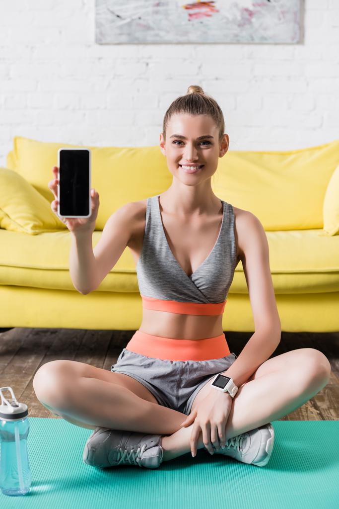 Deportiva positiva mostrando smartphone en cámara en colchoneta de fitness en casa  - Foto, Imagen