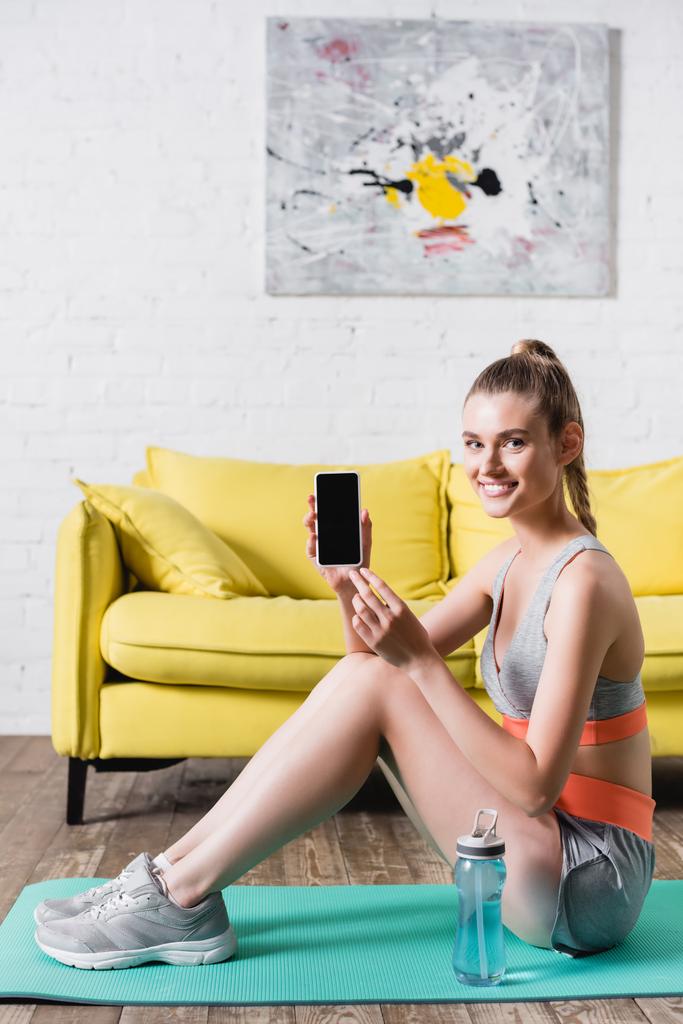 Sportive positive tenant smartphone avec écran blanc près de la bouteille de sport sur tapis de fitness  - Photo, image