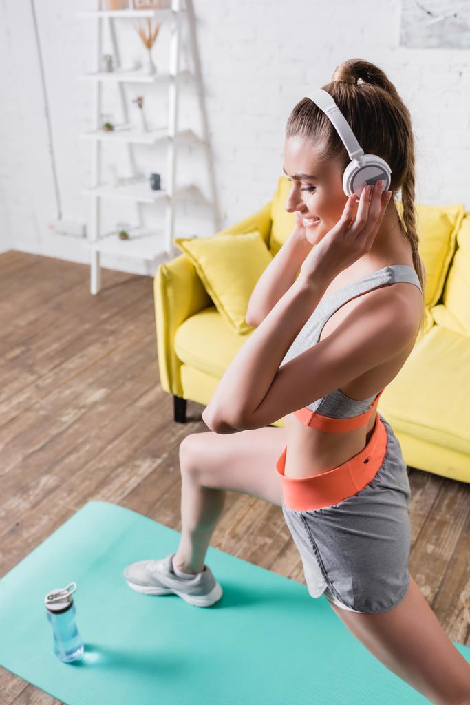 Selective focus of smiling sportswoman in headphones working out on fitness mat in living room  - Photo, Image