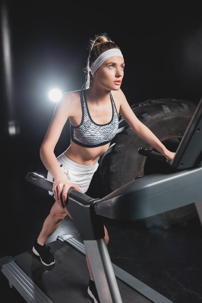 Selective focus of young sportswoman running on treadmill near tire in sports center  - Photo, Image