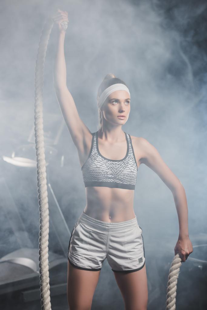 Enfoque selectivo de la joven deportista sosteniendo la cuerda de batalla en el gimnasio con humo  - Foto, imagen