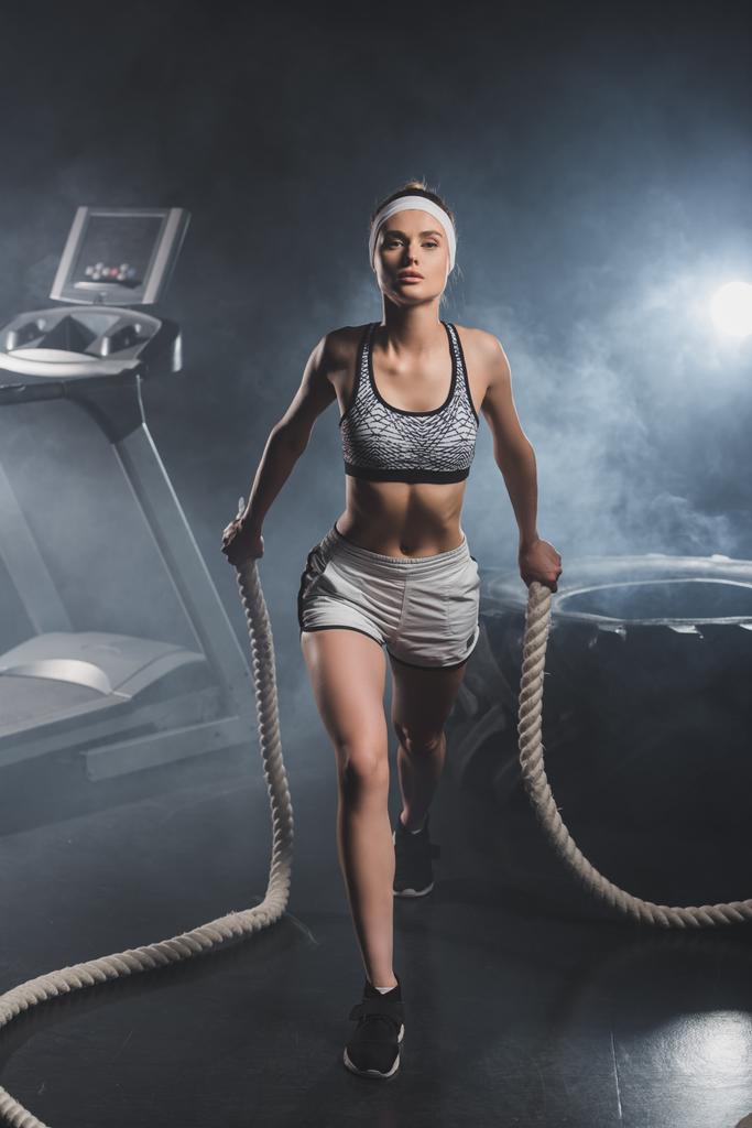 Young sportswoman training with battle rope near treadmill and tire in gym with smoke  - Photo, Image