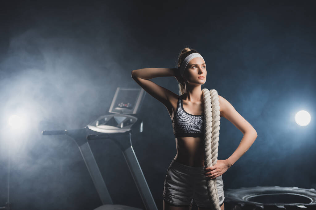 Young sportswoman with battle rope looking away near tire and treadmill in gym with smoke  - Photo, Image