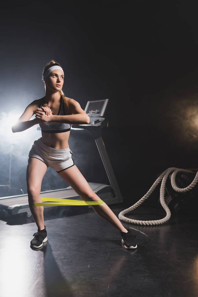 Young sportswoman exercising with resistance band near battle rope and treadmill in gym with smoke  - Photo, Image