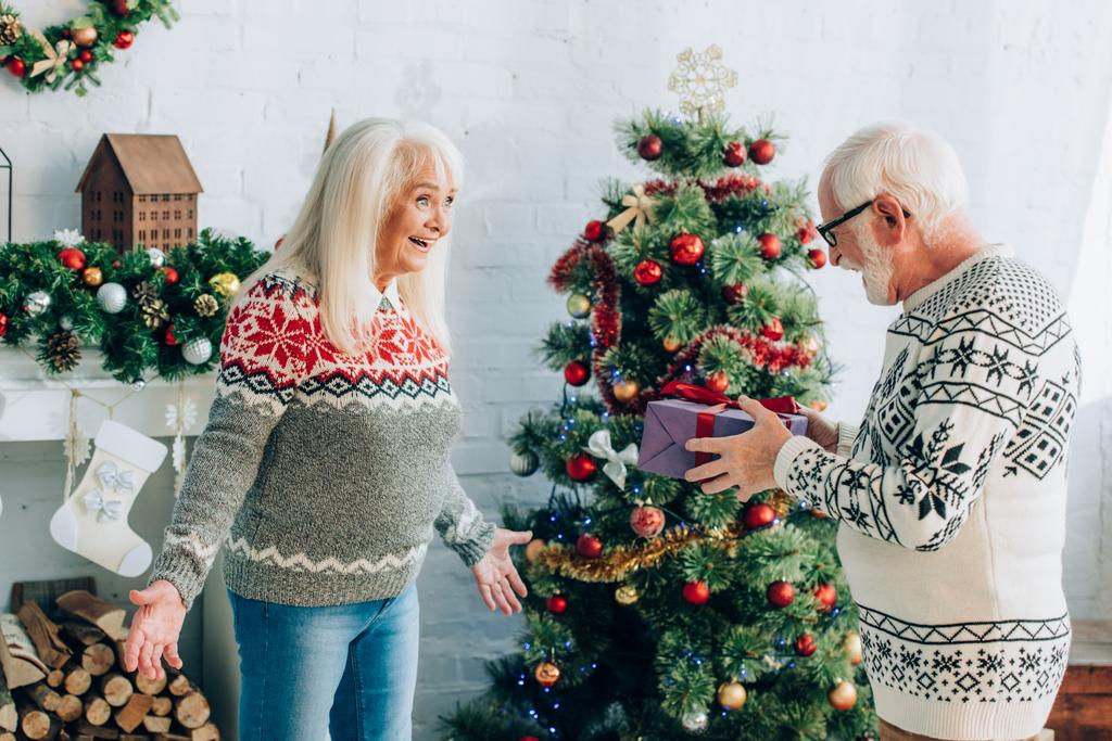 Senior hält Geschenkbox neben aufgeregter Frau, die mit offenen Armen neben Weihnachtsbaum steht - Foto, Bild