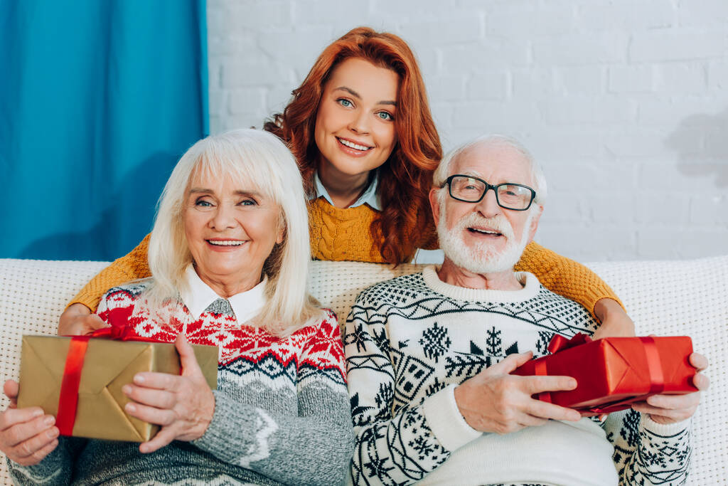 smiling woman standing behind happy senior parents holding gift boxes and smiling at camera - Photo, Image