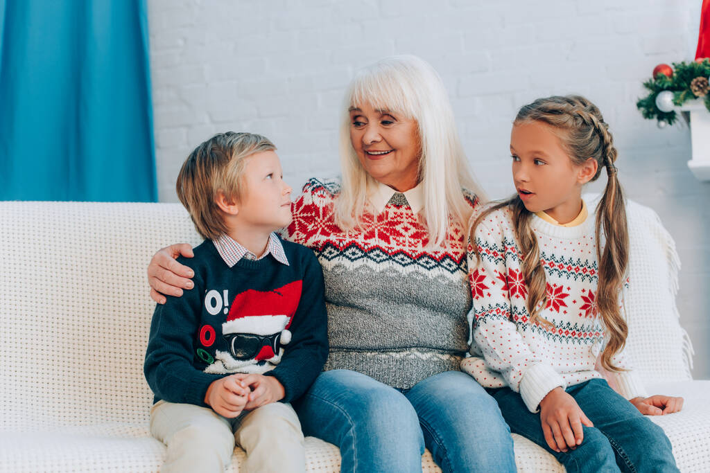 улыбающаяся пожилая женщина разговаривает с внуками, сидя вместе на диване - Фото, изображение