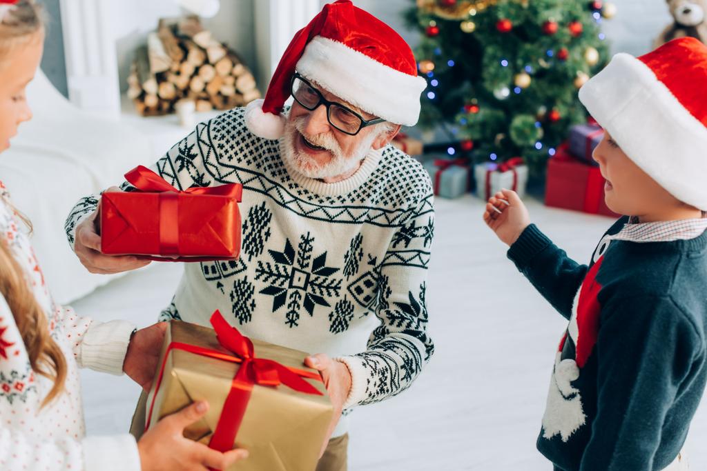 girl and boy presenting gift boxes to happy grandfather in santa hat - Photo, Image