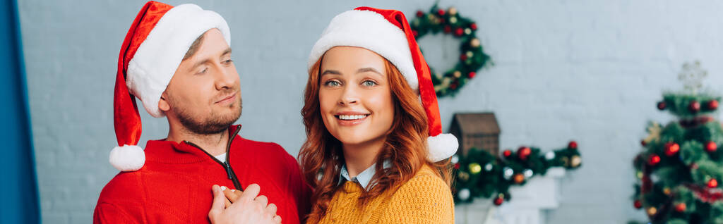 horizontaal beeld van vrouw in santa hoed glimlachen op camera terwijl omarmen echtgenoot op kerst - Foto, afbeelding