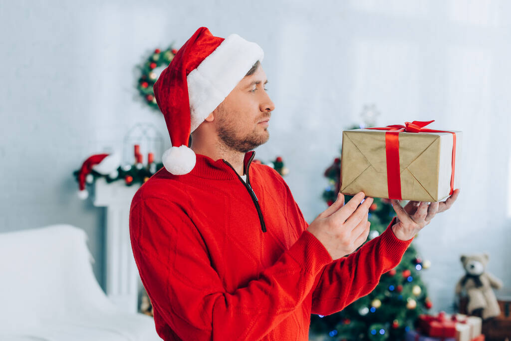 férfi piros pulóverben és Mikulás kalap kezében karácsonyi ajándék doboz - Fotó, kép