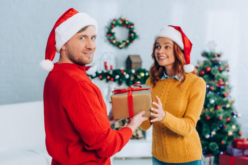 hymyilevä mies santa hattu katselee kameraa esitellessään lahjapakkauksen innoissaan vaimo - Valokuva, kuva