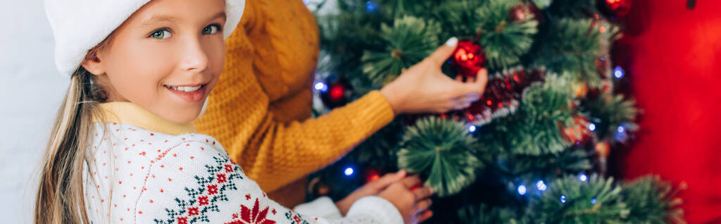cabeçalho do site da menina feliz olhando para a câmera enquanto decora árvore de natal com a família - Foto, Imagem