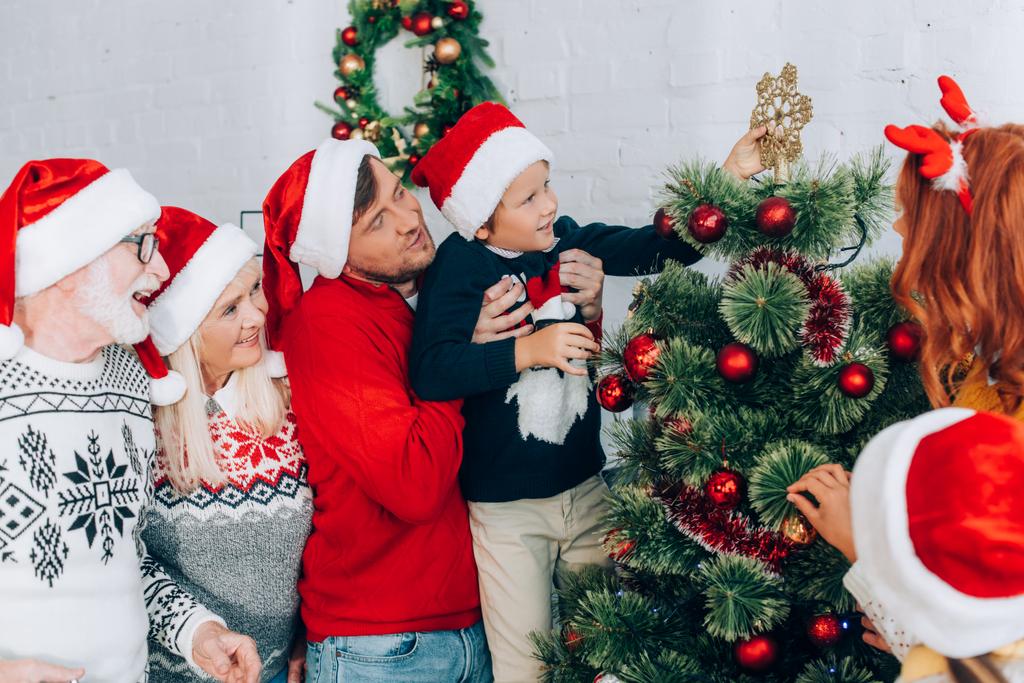 vader houden zoon in de handen, terwijl versieren kerstboom samen met familie - Foto, afbeelding