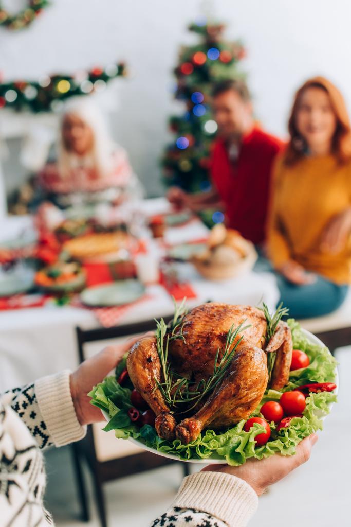 homem sênior segurando delicioso peru assado perto da família sentado na mesa festiva - Foto, Imagem