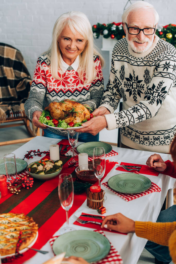 Grand-parents souriants servant la dinde sur la table de fête près de la famille à la maison - Photo, image