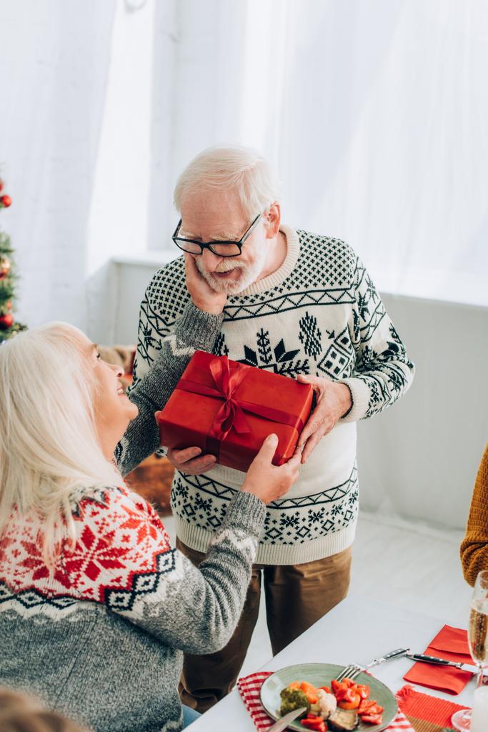 Idősebb nő fogja a kezét a férj arcán, miközben otthon ajándékdobozt visz. - Fotó, kép