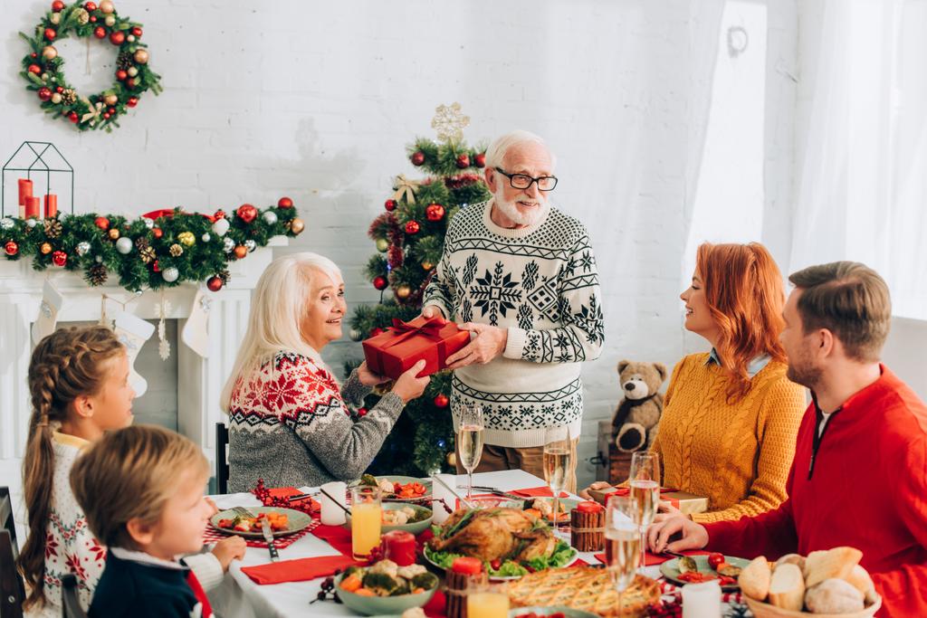Feliz pareja de ancianos sosteniendo caja de regalo cerca de la familia sentada en la mesa festiva - Foto, imagen
