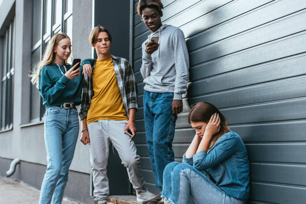 Selective focus of sad girl covering ears near multiethnic teenagers with smartphones and building  - Photo, Image