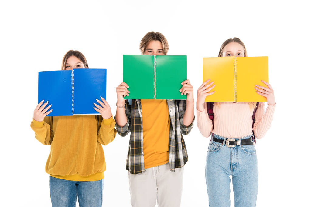 Nastolatki zakrywające twarze książkami do kopiowania i patrzące na kamerę odizolowaną na biało - Zdjęcie, obraz