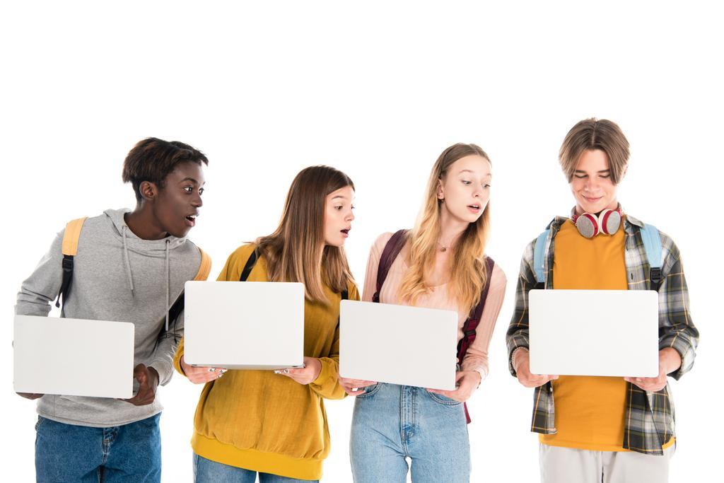 Nadšení multietnické teenageři při pohledu na notebook poblíž přítele s batohem izolované na bílém - Fotografie, Obrázek