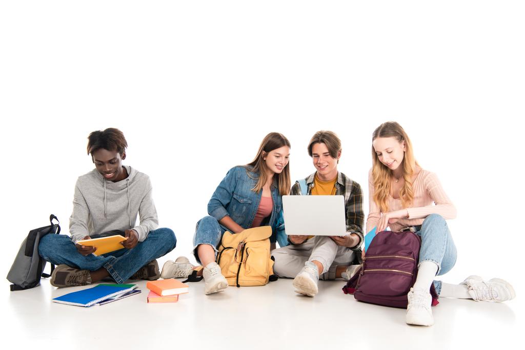 Adolescents multiculturels souriants utilisant un ordinateur portable près des livres et des sacs à dos sur fond blanc - Photo, image