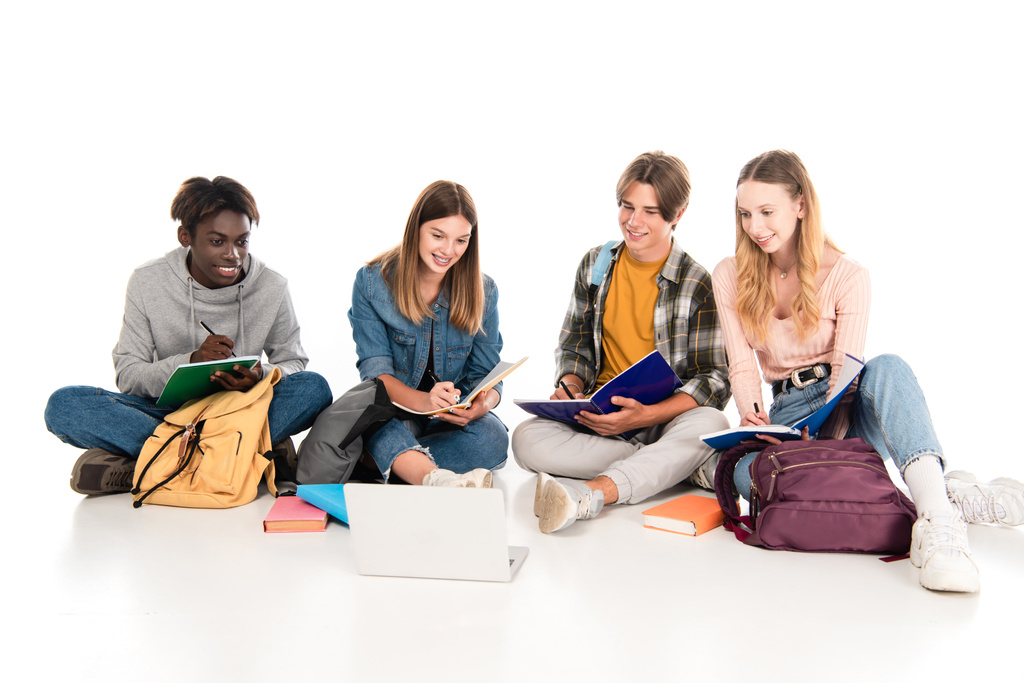 Smiling multicultural teenagers writing on notebooks near laptop on white background - Photo, Image
