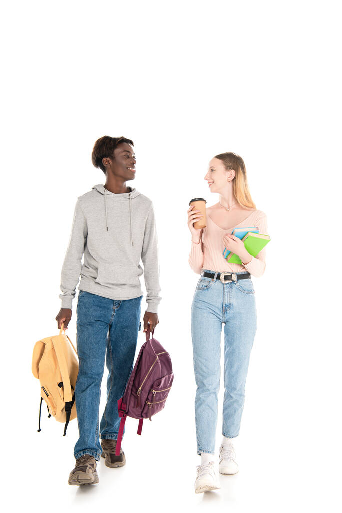 Sonriente adolescente afroamericano sosteniendo mochilas cerca de amigo con café para llevar y libros sobre fondo blanco - Foto, imagen