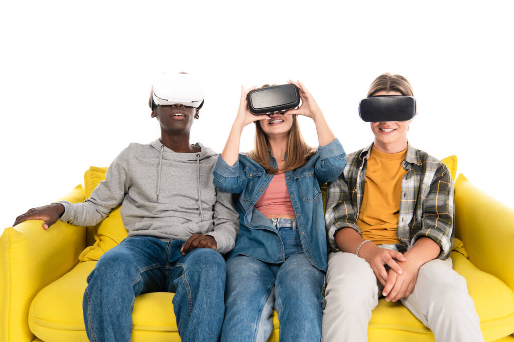Smiling multicultural teenagers in vr headsets sitting on yellow couch isolated on white - Photo, Image