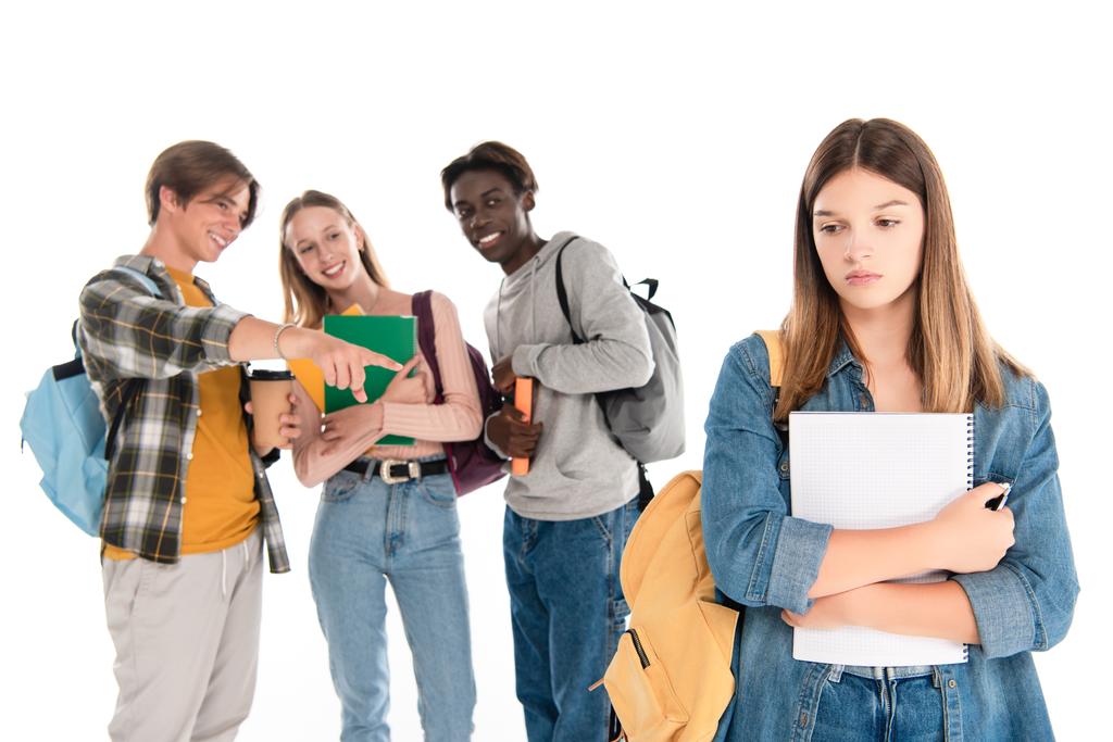 Selektivní zaměření multietnických teenagerů usmívajících se poblíž smutné dívky s notebookem izolovaným na bílém - Fotografie, Obrázek