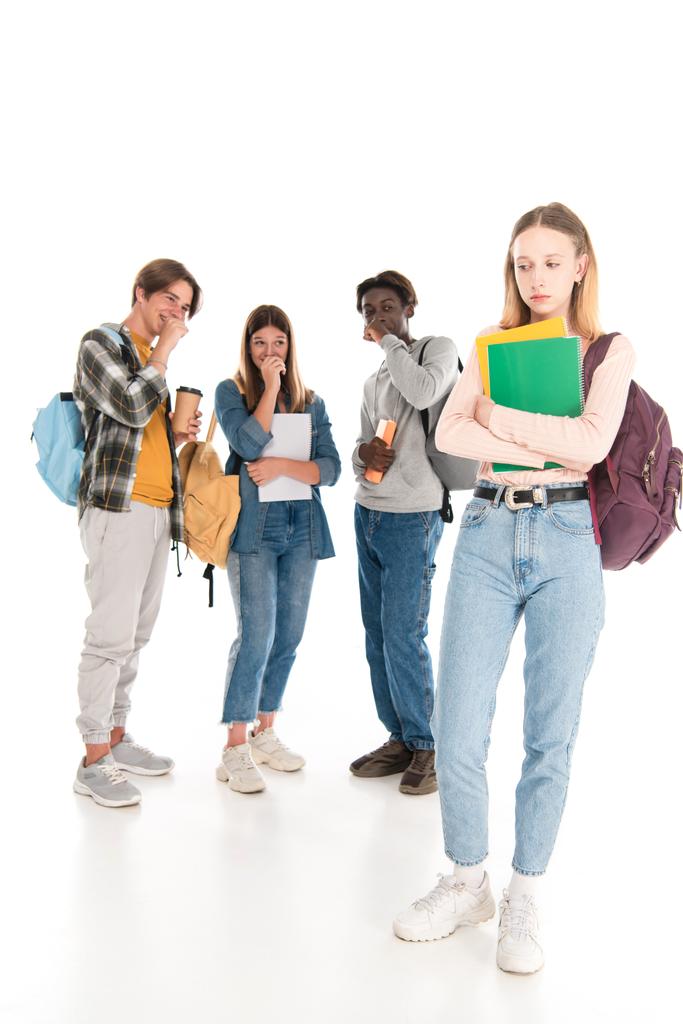 Trauriges Mädchen mit Notizbüchern, die neben lächelnden multiethnischen Teenagern auf weißem Hintergrund stehen - Foto, Bild
