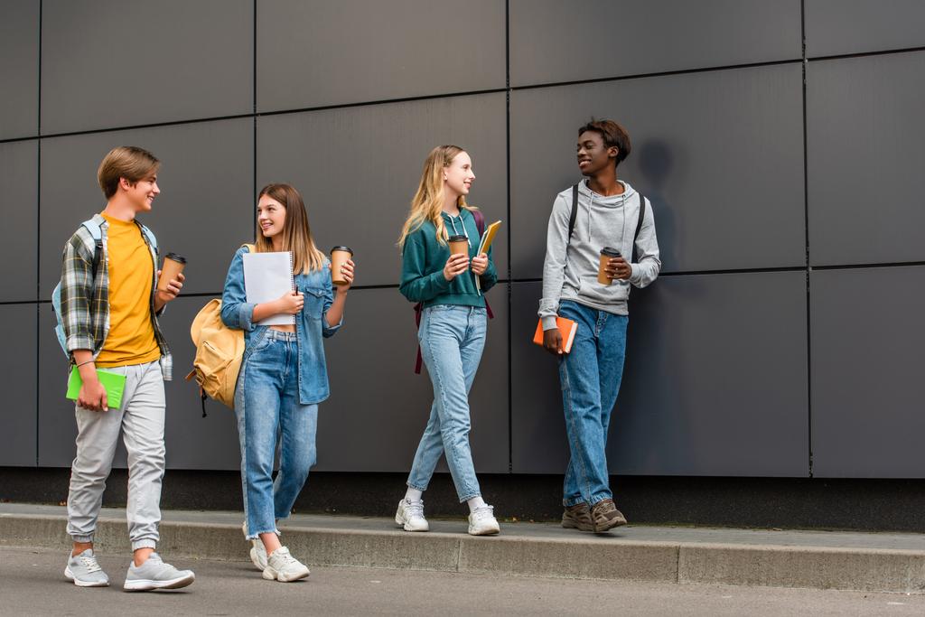 Усміхнені багатоетнічні підлітки з кавою йдуть і блокноти, що йдуть біля будівлі на відкритому повітрі
  - Фото, зображення