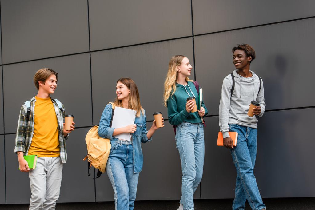 Pozitív többnemzetiségű tinédzserek hátizsákkal és kávéval, akik az épület közelében sétálnak  - Fotó, kép